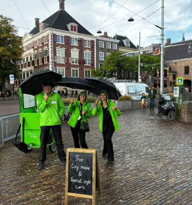 Drie mensen in groene jassen met paraplu's bieden gratis stadskaarten aan.